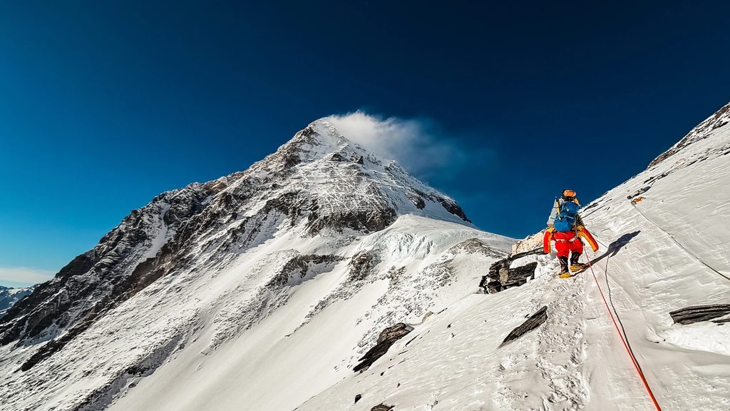 Montanhista em uma encosta de neve e gelo, com o pico do Everest ao fundo, em imagem do projeto Everest Unlimited